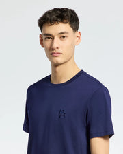 A person with short dark hair, dressed in a Psycho Bunny navy blue MENS NICHOLSON PB CHEST GRAPHIC TEE, looks towards the camera. The pima cotton tee features a subtle embroidered logo on the chest. The background is plain and light-colored.