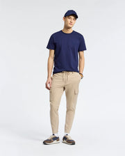 A man in a navy cap and Psycho Bunny MENS NICHOLSON PB CHEST GRAPHIC TEE stands against a plain background in beige jogger pants and sneakers, hands in his pockets beneath the embroidered logo, and wears a watch on his left wrist.