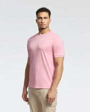 A man with short curly hair wears the Psycho Bunny MENS BRYCE PERFORMANCE TEE in pink, made from a premium Modal-Pima cotton blend, featuring a minimalist logo. Paired with beige pants, the Peruvian-made outfit stands out against a plain white background.