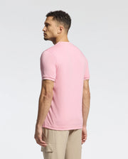 A man with curly hair stands leftward in a pink Psycho Bunny MENS BRYCE PERFORMANCE TEE - B6U490F200, made from a Modal-Pima cotton blend. Paired with beige pants, he poses against a plain white background. This stylish Peruvian-made piece offers both comfort and quality.
