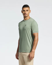 A man with short black hair stands against a plain background, wearing a light green Psycho Bunny MENS BRYCE PERFORMANCE TEE (B6U490F200) made with Peruvian craftsmanship, featuring a small leaf emblem, and beige pants. He looks straight ahead with a neutral expression.