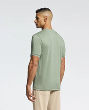 A person with short hair stands sideways, looking away, wearing Psycho Bunny's Men's Bryce Performance Tee in light green, crafted from soft Pima cotton, paired with beige pants. The plain background subtly highlights the elegance of Peruvian craftsmanship.