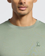 Someone wearing the green Psycho Bunny MENS BRYCE PERFORMANCE TEE, made from soft Peruvian Pima cotton, featuring a small embroidered bunny and crossed bones emblem on the chest, with their face partially visible against a plain background.