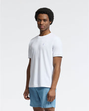 A person wearing a Psycho Bunny MENS BRYCE PERFORMANCE TEE - B6U490F200 and blue shorts stands against a light gray background. Their afro hairstyle frames their face as they look directly at the camera, exuding a relaxed yet stylish vibe in the Modal-Pima cotton blend tee.