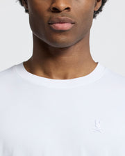 A close-up shows a person wearing the Psycho Bunny MENS BRYCE PERFORMANCE TEE - B6U490F200, a slim-fit, white T-shirt crafted from a luxurious Modal-Pima cotton blend, adorned with an embroidered rabbit and crossed bones logo. The plain white background accentuates the exquisite design.