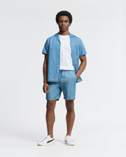 Against a light background, a person wears the Psycho Bunny MENS BRYCE PERFORMANCE TEE - B6U490F200, layered under a light blue slim-fit Modal-Pima cotton blend button-up shirt and matching shorts. They complete the look with white sneakers featuring black accents and stand with one hand in their pocket.