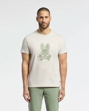 A man in a MENS SYDNEY GRAPHIC TEE - B6U508F200 by Psycho Bunny, featuring a Bunny Graphic with skull and crossbones, stands against a plain white background. He has short hair, a beard, and wears light green pants.