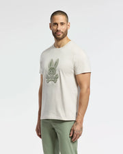 A bearded man stands against a plain white background, wearing a light-colored MENS SYDNEY GRAPHIC TEE - B6U508F200 by Psycho Bunny, featuring a stylized crossbones bunny graphic. He pairs it with light green pants.