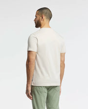 A man stands with his back to the camera, wearing a Psycho Bunny MENS SYDNEY GRAPHIC TEE (B6U508F200) in light beige Pima cotton featuring a subtle bunny graphic. His sage green pants enhance the minimalist vibe while he poses against a plain white background, emphasizing the clean aesthetic.