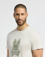 A person with a buzz haircut and beard wears the Psycho Bunny MENS SYDNEY GRAPHIC TEE - B6U508F200, a cream-colored Pima cotton shirt featuring a stylized bunny skull and crossbones graphic, set against a plain white background.