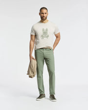 A man with short hair and a beard stands against a plain background, wearing a beige Psycho Bunny Pima cotton MENS SYDNEY GRAPHIC TEE with a bunny graphic, green pants, and dark sneakers. He holds a matching beige jacket in his left hand while casually slipping his right hand into his pocket.