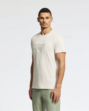 A man with short dark hair stands slightly angled, wearing a light beige Psycho Bunny MENS SYDNEY WORDMARK GRAPHIC TEE (B6U510F200) and light green pants against a plain white background.