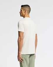 A man faces away from the camera, wearing a Psycho Bunny MENS SYDNEY WORDMARK GRAPHIC TEE (B6U510F200) in light beige and light green pants against a plain white background.