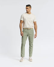A man stands against a plain white background, wearing the Psycho Bunny MENS SYDNEY WORDMARK GRAPHIC TEE in beige, paired with light sage green pants, a beige belt, and beige-and-white sneakers. He faces sideways with a relaxed pose.