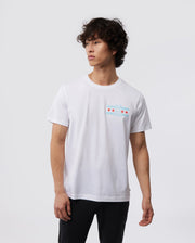 Amidst a simple backdrop, an individual with curly hair wears the Psycho Bunny MENS CHICAGO CITY TEE - B6U551W1PC, made from soft Pima cotton. The tee sports a subtle chest graphic of red stars, and they stand calmly with arms relaxed.