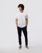 A person with curly hair stands casually against a plain background, wearing a Psycho Bunny MENS CHICAGO CITY TEE - B6U551W1PC, paired with black pants and classic black and white sneakers. Their hands rest in their pockets as they gaze to the side.