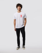 A person with curly hair is standing against a plain background, wearing a Psycho Bunny MENS NEW YORK TEE - B6U554W1PC made of soft Pima cotton jersey. The white shirt features a red bunny logo graphic on the chest. They are also dressed in black pants and black and white sneakers, with a neutral expression and their hands loosely by their sides.