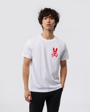 A person with short curly hair is wearing a white Pima cotton jersey T-shirt featuring a red bunny logo with crossed bones beneath it, the MENS NEW YORK TEE - B6U554W1PC from Psycho Bunny. The person is also wearing dark pants and standing against a plain light gray background.