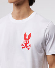 A person wearing a white Psycho Bunny MENS NEW YORK TEE - B6U554W1PC made of soft Pima cotton jersey. The red graphic on the chest features a bunny logo with ears shaped like the letters "NY" and crossed bones underneath. The photo shows the chest and neck area, but the person's face is not visible.