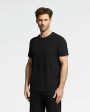 A man with short brown hair is wearing Psycho Bunny's plain black Taylor Pique Tee (B6U579F200) and black pants. He stands against a light gray background, smiling slightly, his outfit subtly elevated by the tee's lightweight piqué texture.