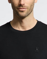 A close-up of a man in the MENS TAYLOR PIQUE TEE by Psycho Bunny, featuring lightweight piqué fabric and an embroidered chest Bunny logo with crossed ears on the left. The white background highlights his lower face and upper torso.