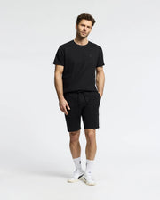 Against a white backdrop, a man wears the MENS TAYLOR PIQUE TEE by Psycho Bunny in black Pima cotton, paired with black shorts. He has his hands in his pockets and complements the look with white socks, sneakers, and a subtle embroidered Bunny on the chest for elegance.