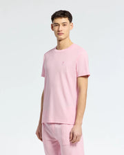 A young man faces the camera with a neutral expression, wearing the Psycho Bunny MENS TAYLOR PIQUE TEE (B6U579F200) in light pink piqué fabric, paired with matching pink pants against a plain light background.