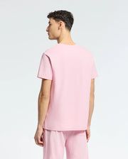 A young man with short curly hair stands facing away, wearing the Psycho Bunny MENS TAYLOR PIQUE TEE (B6U579F200) in light pink piqué fabric, paired with matching pink shorts against a light background.
