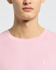 A person wears the Psycho Bunny MENS TAYLOR PIQUE TEE (B6U579F200), a light pink crew neck made from lightweight piqué, featuring a small white embroidered Bunny and crossed bones logo on the left chest. Only their lower face, neck, and upper torso are shown.