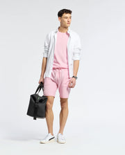 A young man stands against a plain background, wearing Psycho Bunny’s MENS TAYLOR PIQUE TEE (B6U579F200) in light pink with matching shorts, a white open button-down shirt with an embroidered Bunny, white sneakers, and holding a black duffel bag.