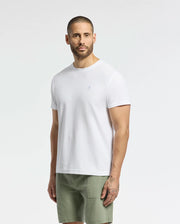 A man with a shaved head, beard, and tattoos on his left hand wears a Psycho Bunny MENS TAYLOR PIQUE TEE (B6U579F200), made from lightweight Pima cotton in regular fit, paired with olive green shorts. He stands against a plain white background, gazing forward with a neutral expression.