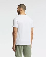 A man stands against a white background, facing away, wearing the Psycho Bunny MENS TAYLOR PIQUE TEE in plain white and light green shorts. The tee is expertly crafted from lightweight Pima cotton.