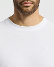 Close-up of a man wearing the Psycho Bunny MENS TAYLOR PIQUE TEE - B6U579F200, a regular-fit white crew-neck shirt in lightweight Pima cotton, featuring a small embroidered Bunny and crossbones emblem on the left chest, focusing on the upper torso and neckline.