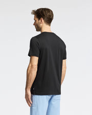 A man with short brown hair stands with his back to the camera, wearing a Psycho Bunny MENS ZEALAND GRAPHIC TEE (B6U604F200) and light blue pants against a white background.