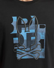 The Psycho Bunny MENS ZEALAND GRAPHIC TEE (B6U604F200) is a black tee with a blue checkered background and a black outlined bunny head above crossed bones, creating a playful skull and crossbones look.