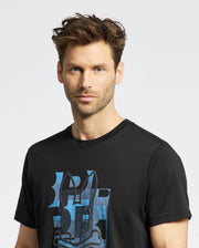 A man with tousled brown hair and light stubble wears the Psycho Bunny MENS ZEALAND GRAPHIC TEE—B6U604F200, a black shirt featuring a blue stylized bunny face. He stands against a white background, looking directly at the camera.