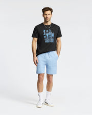 A man with short brown hair and a light beard stands against a white background, wearing the Psycho Bunny MENS ZEALAND GRAPHIC TEE (B6U604F200), light blue drawstring shorts, white socks, and white sneakers with hands in his pockets.