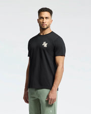 A man with short curly hair is wearing a Psycho Bunny MENS ELLIOT FRONT BACK GRAPHIC TEE in black, featuring a small white logo on the chest. Crafted from Pima cotton, it pairs perfectly with his light green shorts against a plain white background.