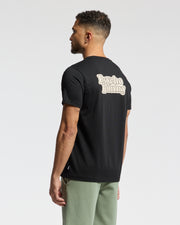 Short-haired man sports a black "MENS ELLIOT FRONT BACK GRAPHIC TEE - B6U605F200" by Psycho Bunny, featuring a back print. Styled with light green pants, the regular fit outfit is crafted from soft Pima cotton. Set against a plain white background, he gazes to the side.