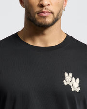 A man wears the Psycho Bunny MENS ELLIOT FRONT BACK GRAPHIC TEE (B6U605F200), a black, regular fit Pima cotton T-shirt. It features a small graphic of two cartoon-style rabbit skulls with crossed bones on the left chest, adding a playful yet edgy flair to any wardrobe.
