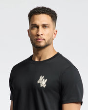A man with curly hair and an earring models the Psycho Bunny Men's Elliot Front Back Graphic Tee (B6U605F200), a soft Pima cotton black T-shirt featuring a stylish rabbit print on the chest. He stands against a plain white background, looking directly at the camera.