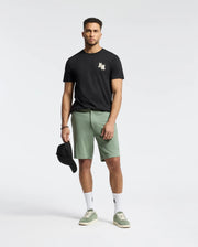A man in a Psycho Bunny regular fit MENS ELLIOT FRONT BACK GRAPHIC TEE - B6U605F200 and green shorts stands against a plain background, holding a black cap in his right hand, wearing white socks and green shoes.
