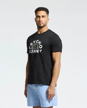 A man in a neutral pose wears the Psycho Bunny MENS GOLDIE GRAPHIC TEE - B6U606F200, crafted from premium Pima cotton, featuring white text and a subtle bunny graphic. He pairs it with light blue shorts against a plain background.