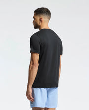 A man with short curly hair, facing away, dons a black Psycho Bunny Men's Goldie Graphic Tee (B6U606F200) made from Pima cotton paired with light blue shorts against a plain, light background.