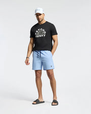 A man stands against a plain background wearing a Psycho Bunny MENS GOLDIE GRAPHIC TEE - B6U606F200, light blue shorts, black slides, and a white cap.