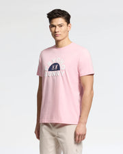 Someone is sporting a light pink Psycho Bunny MENS GOLDIE GRAPHIC TEE made from Peruvian Pima cotton, featuring "Psycho Bunny" and a bunny graphic on the front, paired with light-colored pants against a plain backdrop.