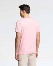 A person with short dark hair wears a Psycho Bunny MENS GOLDIE GRAPHIC TEE - B6U606F200 made of soft Peruvian Pima cotton in light pink, paired with beige shorts, standing and facing away against a plain white background.