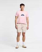 A man is wearing a pink Psycho Bunny MENS GOLDIE GRAPHIC TEE with a bunny design, paired with beige shorts, white socks, and beige sneakers against a plain white background.