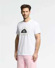 Someone is wearing the MENS GOLDIE GRAPHIC TEE - B6U606F200 by Psycho Bunny, a soft Pima cotton white T-shirt with the iconic bunny and skull print, paired with light pink shorts on a plain white background.