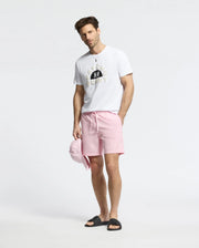 A man stands against a plain background wearing an MENS GOLDIE GRAPHIC TEE from Psycho Bunny, paired with pale pink shorts and black sandals. He holds a light pink cap in one hand, showcasing the soft luxury of Peruvian cotton.